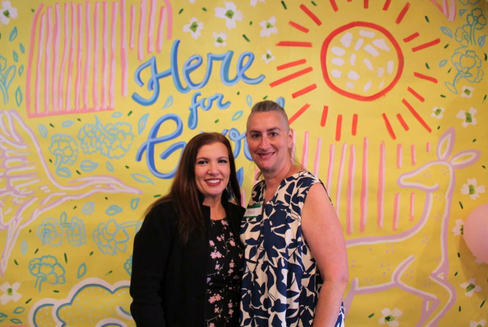 Two women smiling and standing close together in front of a bright yellow wall with colorful drawings and the words "Here for Good" on it. The wall features a sun, a deer, and various decorative elements.