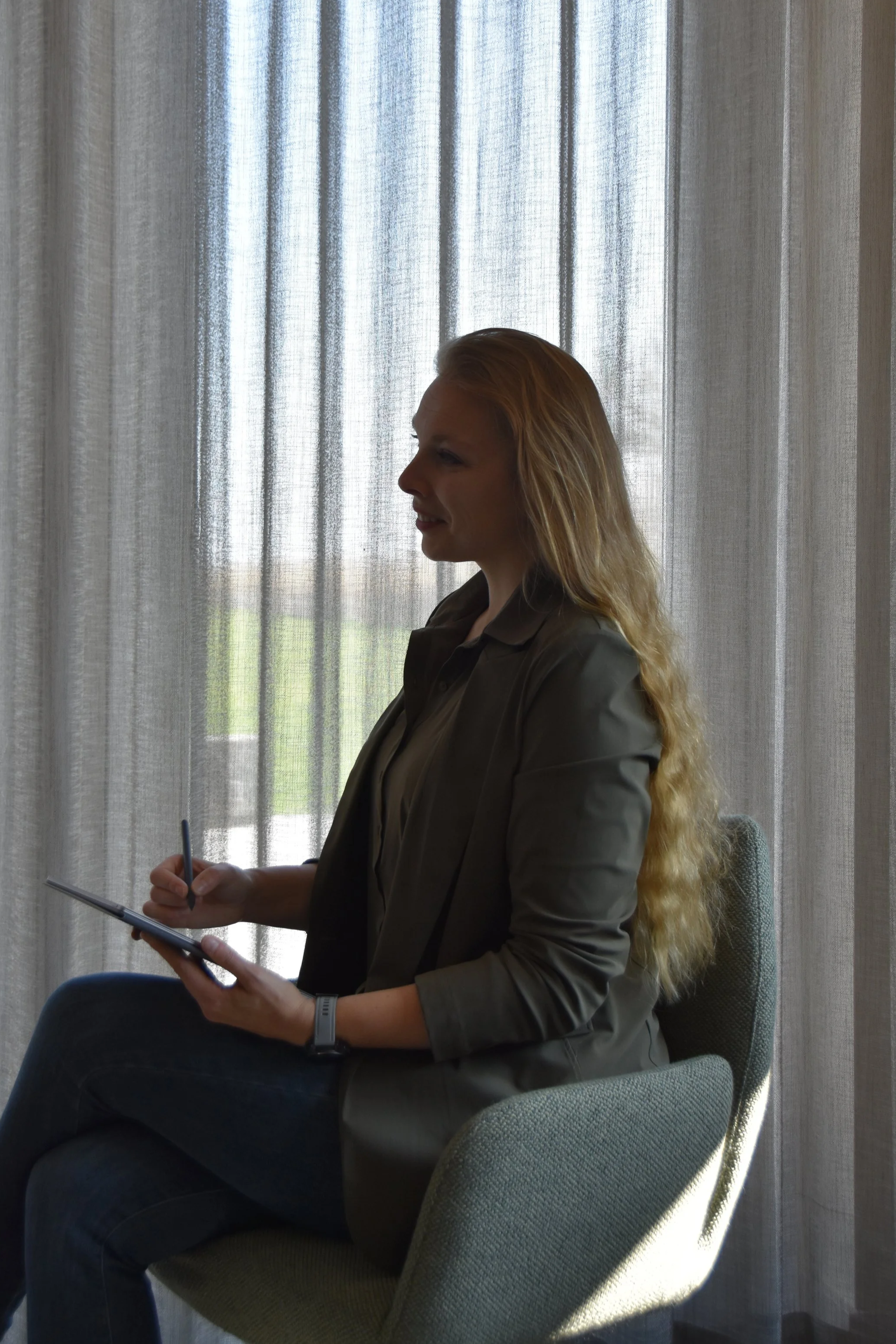 A woman with long, wavy red hair sitting on a green upholstered chair in front of sheer curtains, holding a tablet and stylus, with natural light coming through the curtains.