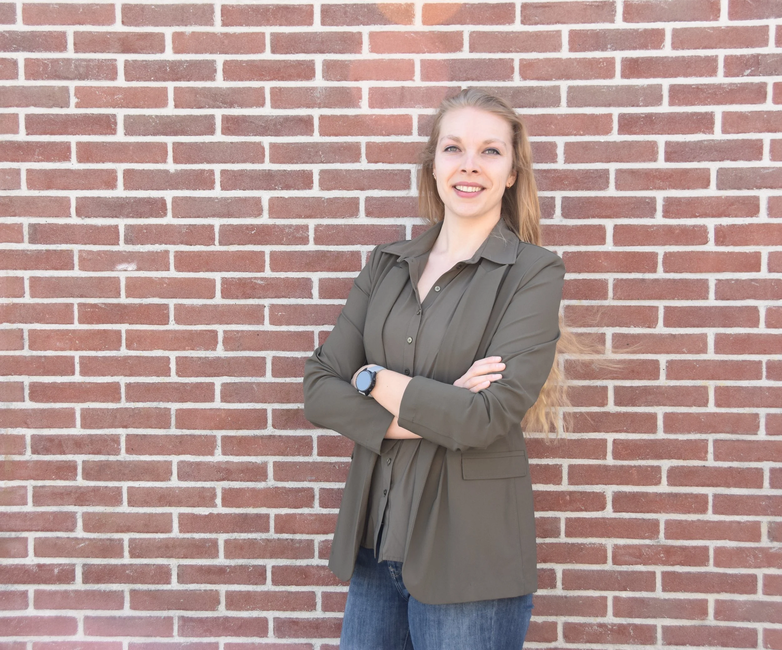 A woman with long, light brown hair standing with her arms crossed in front of a red brick wall, wearing a green button-up shirt and a dark wristwatch, smiling at the camera.