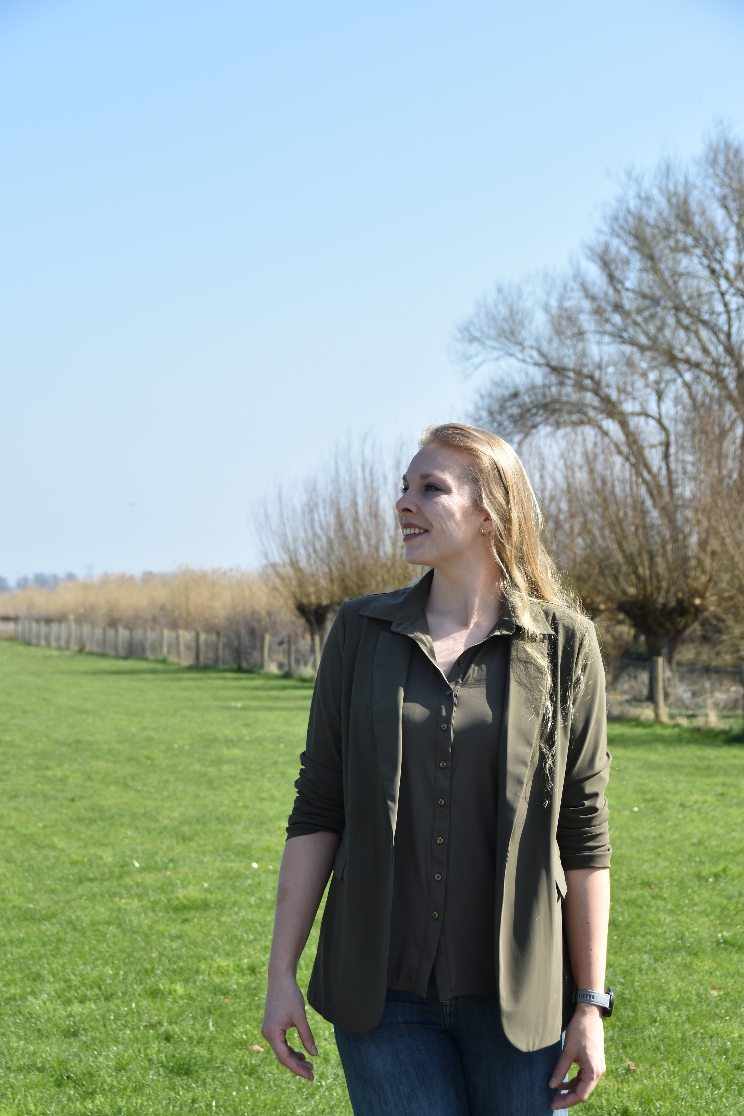 A woman smiling outdoors in a grassy field with trees in the background, wearing a dark green shirt and jacket.