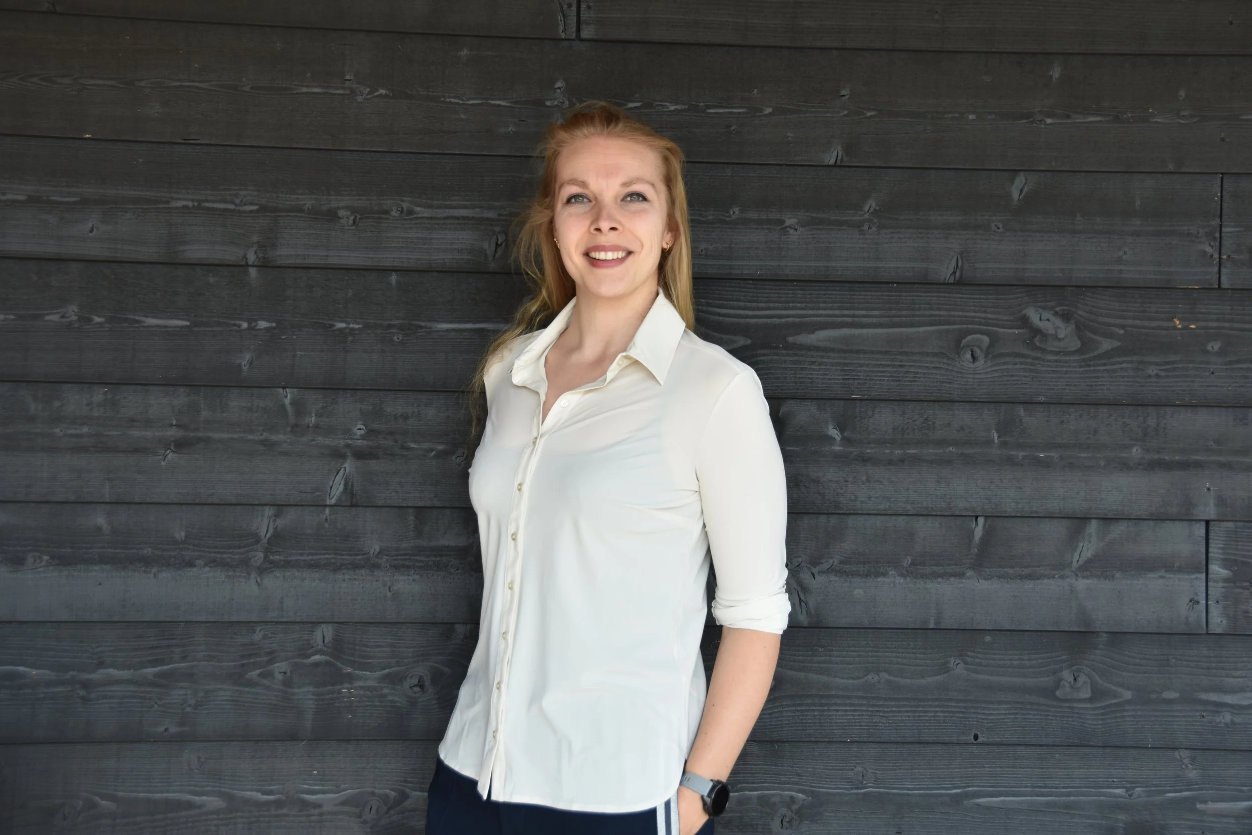 A woman with red hair standing against a dark wooden wall, wearing a white shirt and a smartwatch.