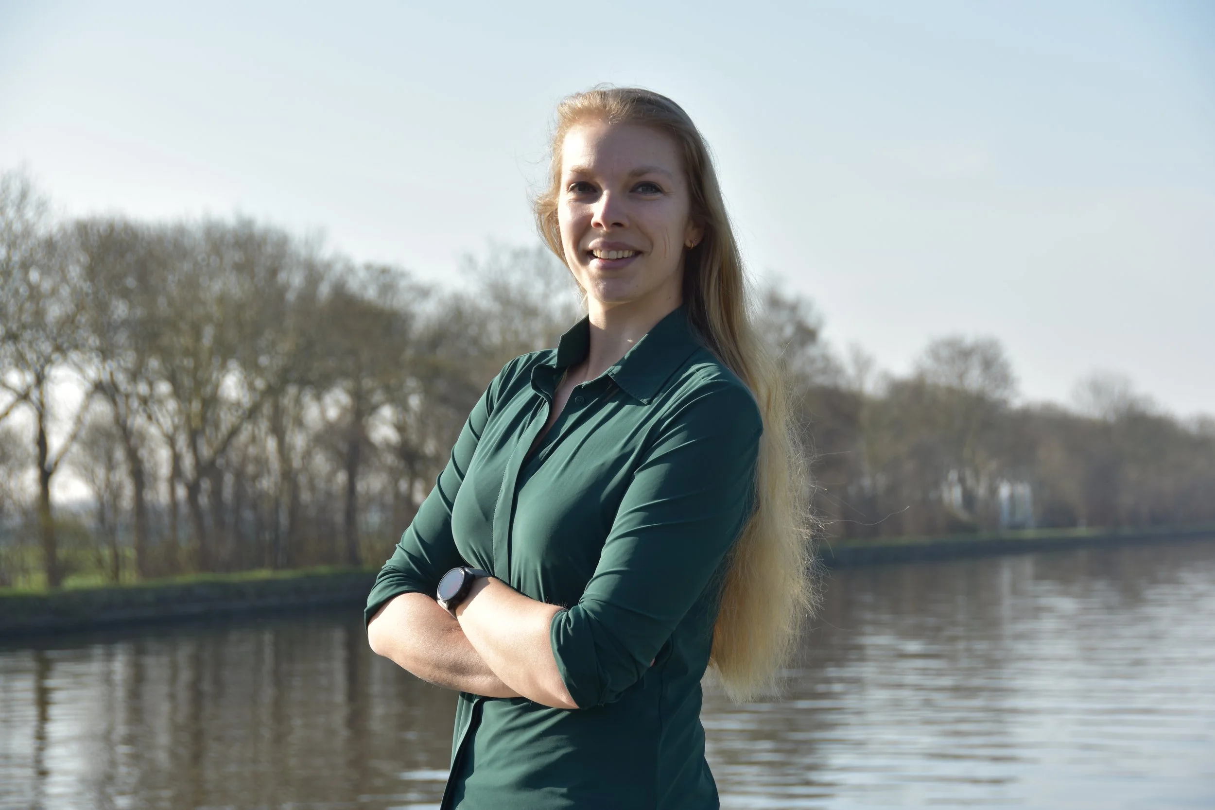 A woman with long blonde hair standing near a river with trees in the background, crossing her arms and smiling at the camera on a sunny day.