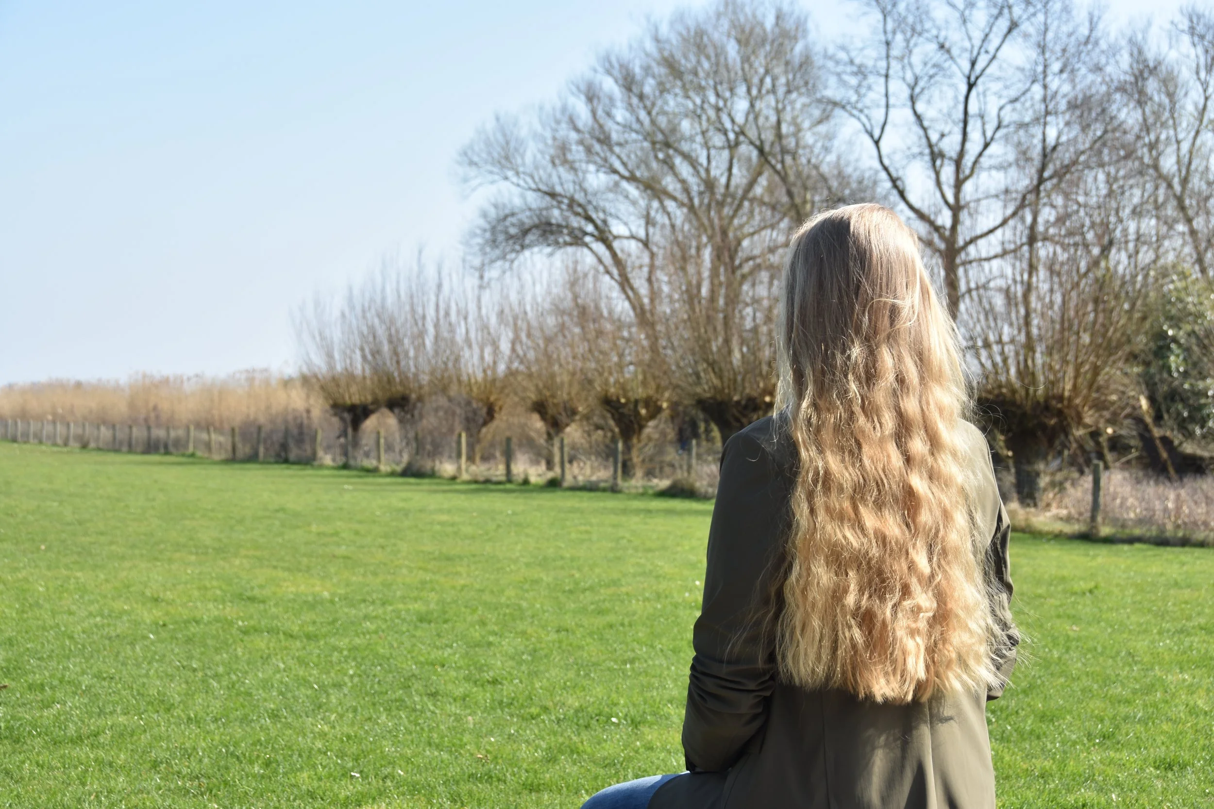 A woman with long, wavy blonde hair sitting on green grass in a park, facing away from the camera, with leafless trees in the background under a clear blue sky.
