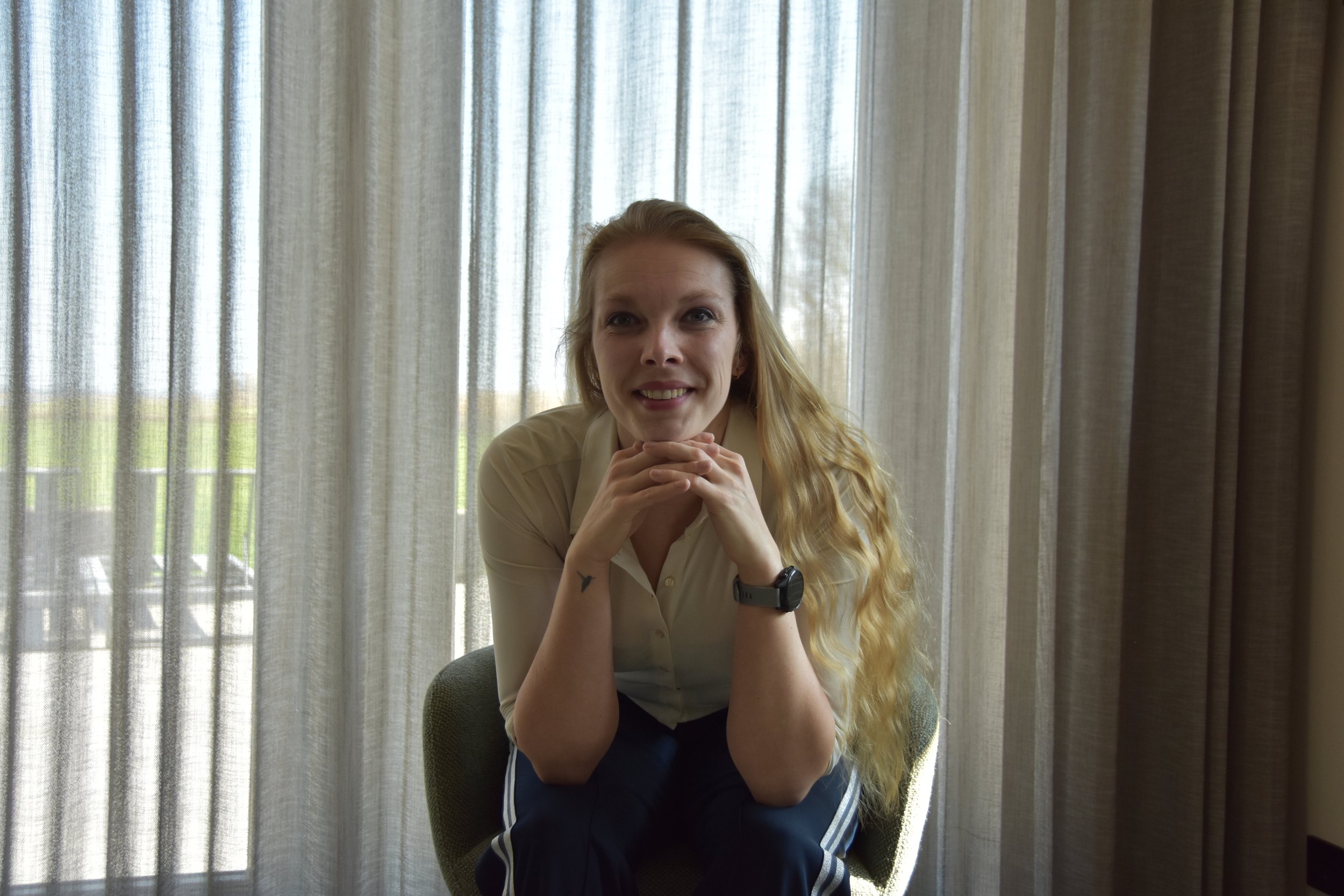 A young woman with long, curly blonde hair sitting in an armchair near large windows with sheer curtains, smiling and looking at the camera.