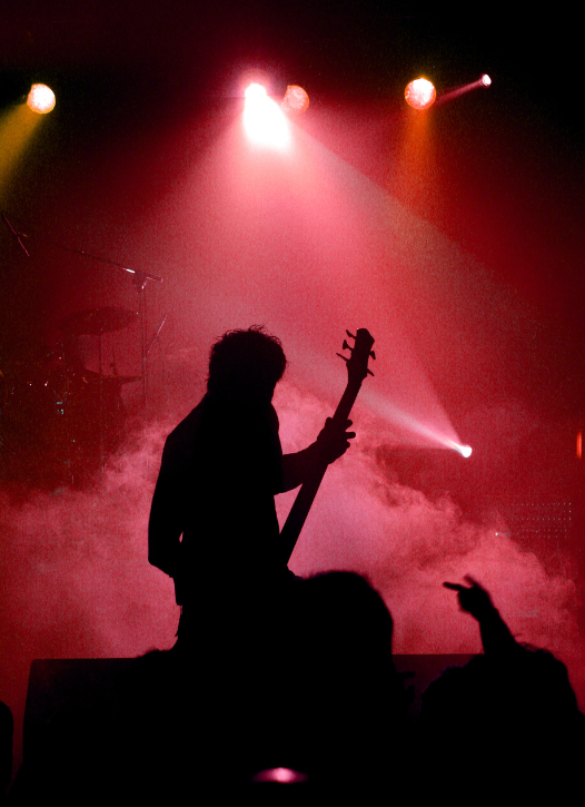 Silhouette of a musician playing electric guitar on stage with colorful stage lights and smoke, audience member raising a finger.