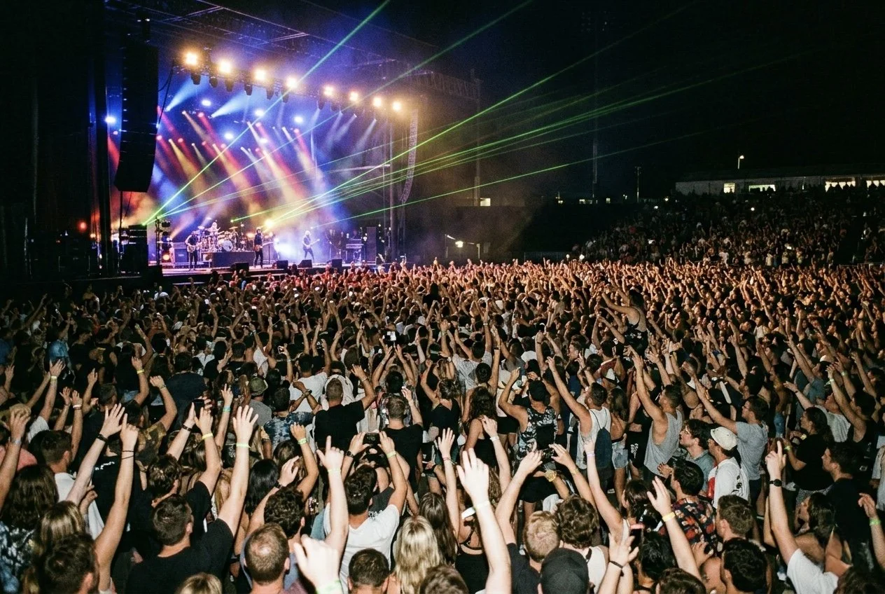 Crowd of concert attendees raising their hands while watching a band on stage with colorful lights and laser beams at night.