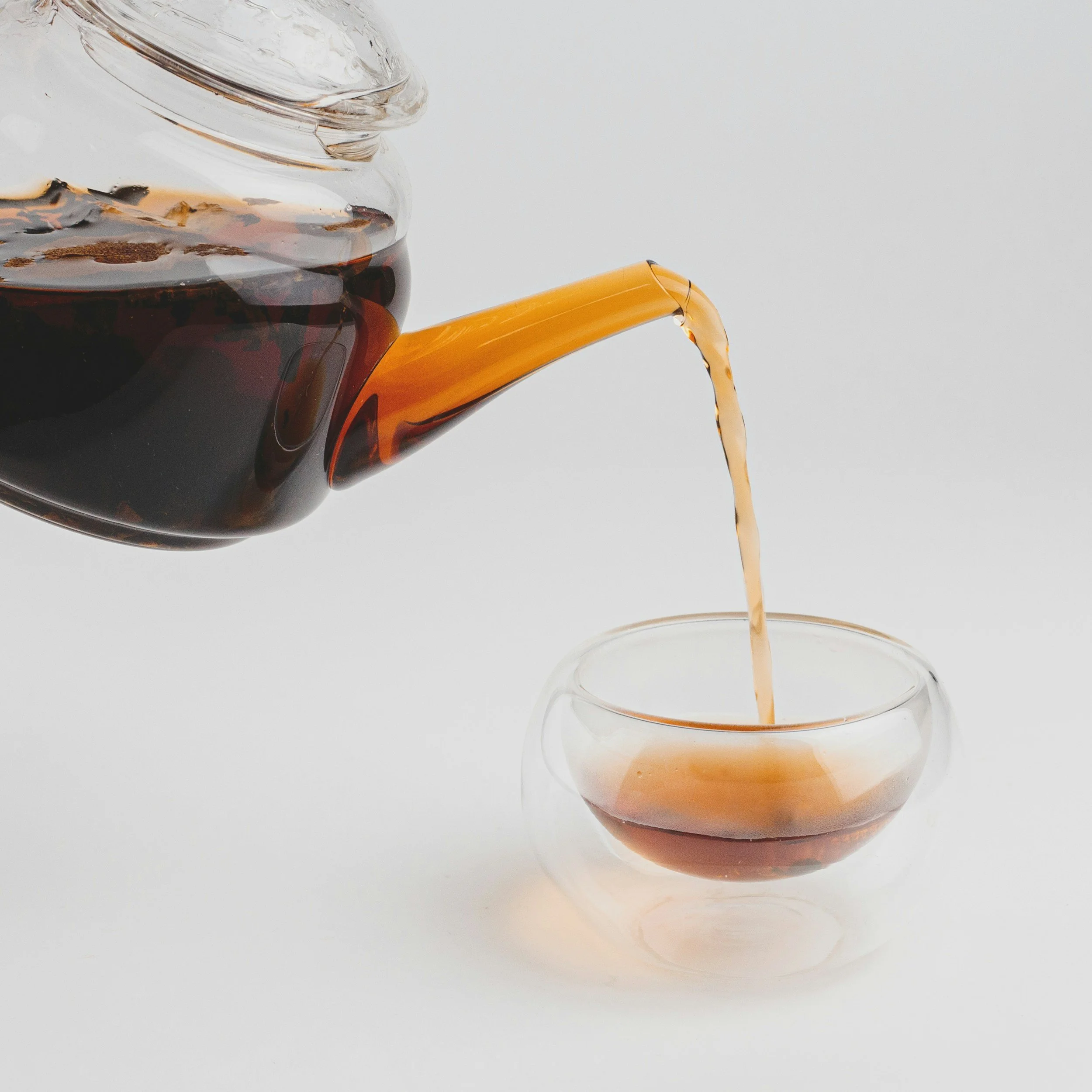 A glass teapot pouring dark tea into a small glass cup on a white surface.