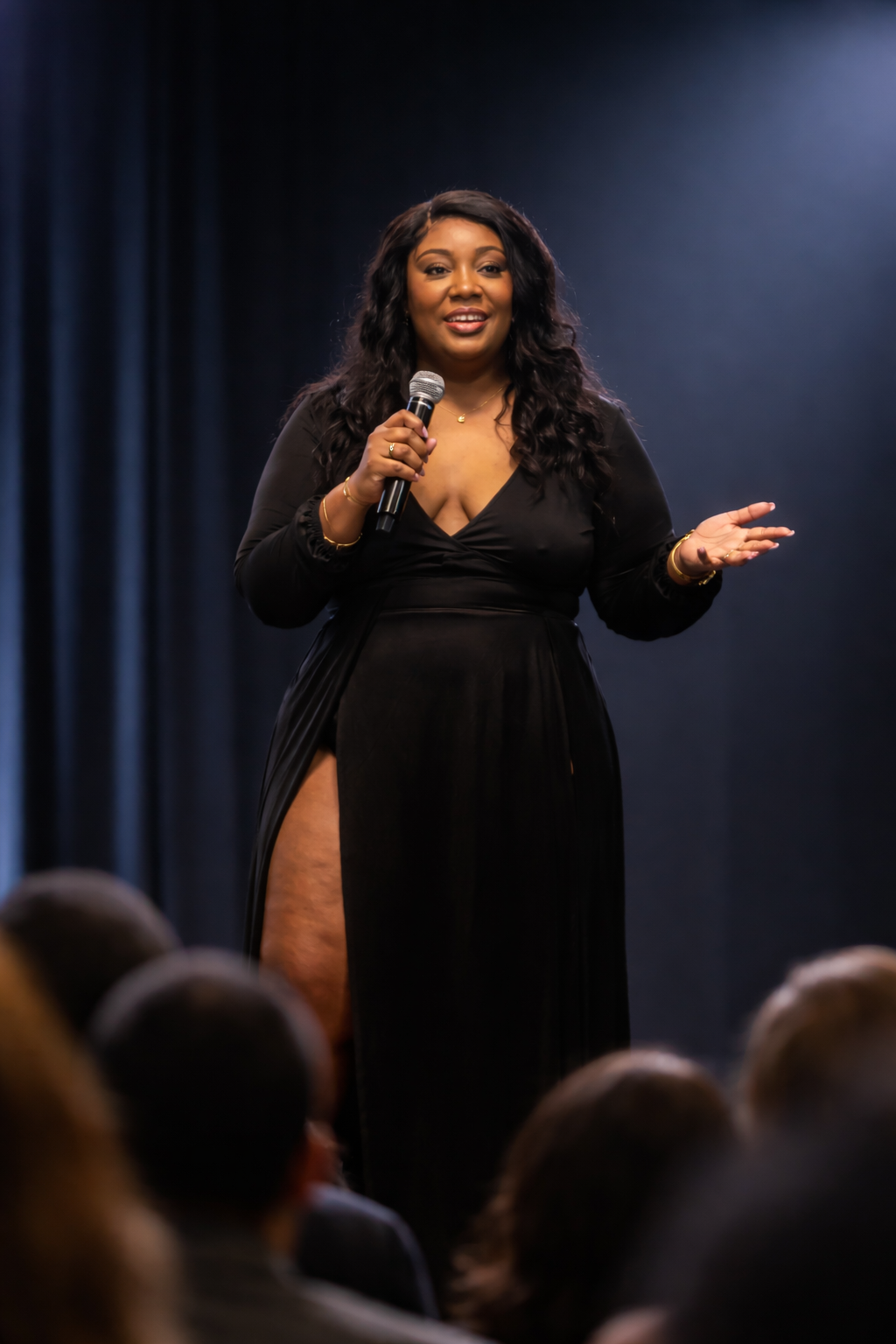 A woman in a black dress holding a microphone, standing on stage in front of an audience, gesturing with her left hand.