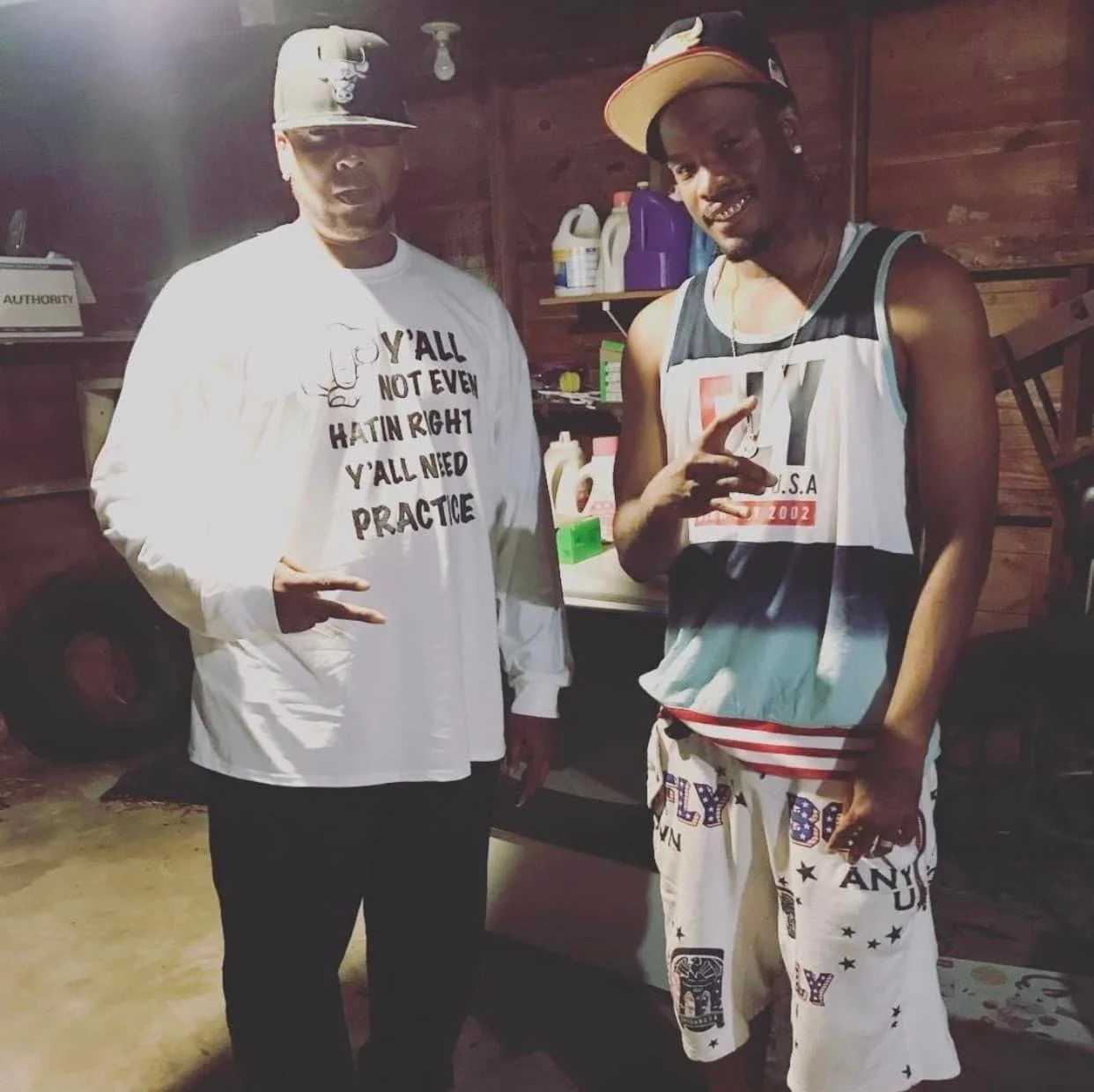 Two young men stand indoors against a wooden wall, making hand gestures. The man on the left wears a white long-sleeve shirt and a beige baseball cap, while the man on the right wears a sleeveless football jersey and patterned shorts, accessorized wi