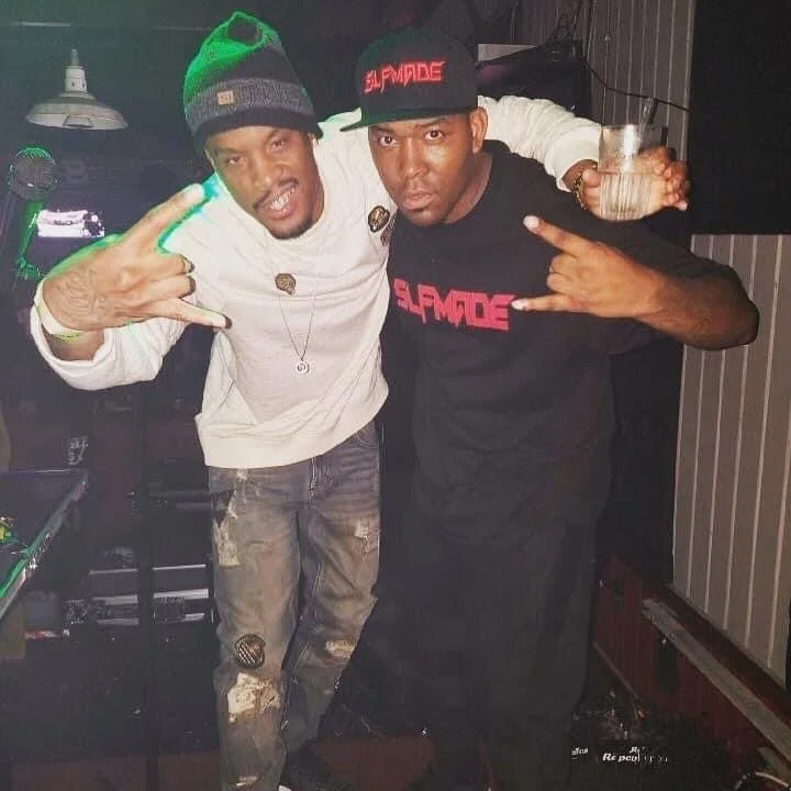 Two men posing in a dimly lit nightclub or bar, making peace signs with their hands. The man on the left wears a striped beanie, a white long-sleeve shirt, and distressed jeans. The man on the right wears a black cap with red writing and a black T-sh