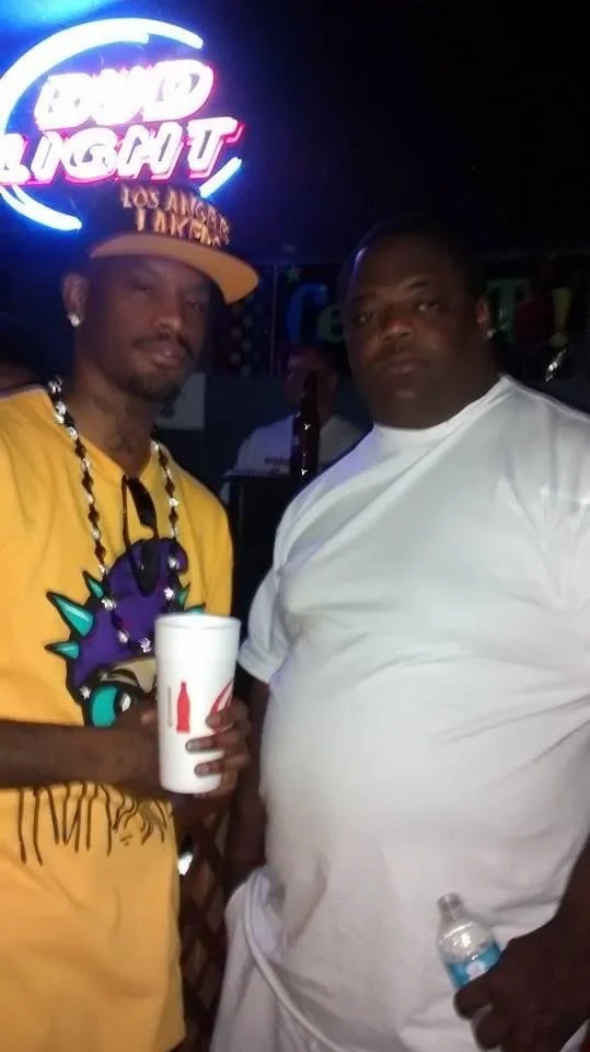 Two men in a bar with a neon Bud Light sign in the background. One man wears a yellow shirt, a hat with the text "Los Angeles" and a beaded necklace, holding a white cup. The other man is dressed in a white shirt, holding a water bottle.