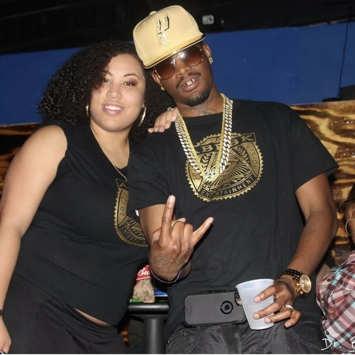Two people at a party, one woman and one man, smiling and posing for the camera. The woman has curly hair, and the man is wearing a gold cap, sunglasses, and gold chains. The man is making a hand gesture, and both are dressed in black themed clothing