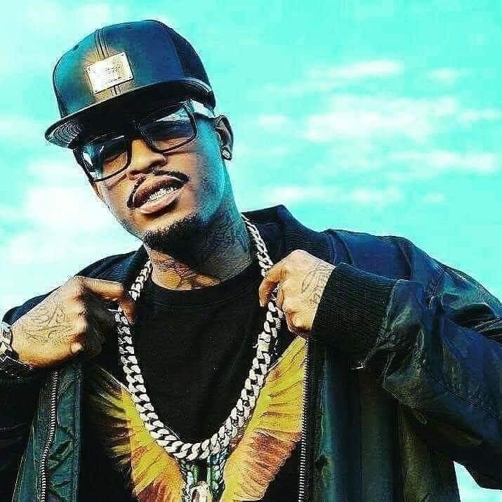 A man wearing a black leather jacket, black shirt with a printed design, black baseball cap, sunglasses, and multiple gold chains, standing outdoors against a bright blue sky.