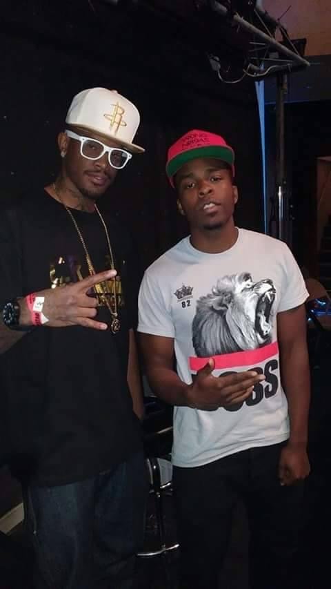 Two young men posing for a photo. The man on the left is wearing a white baseball cap with a gold logo, white glasses, a black T-shirt, and a watch. The man on the right is wearing a white T-shirt with a lion graphic, a red and green baseball cap, an