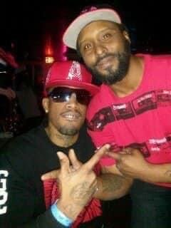 Two men posing together; one wearing sunglasses and a red cap, the other wearing a white and pink cap, both in dark clothing.