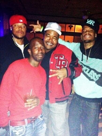 Four men standing together in a dimly lit indoor setting, smiling for the camera, with one holding a drink. They are wearing casual clothing and baseball caps.