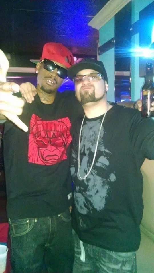 Two men are at a party or club. The man on the left is wearing a red cap, sunglasses, a black T-shirt with a red graphic, and is making a hand gesture. The man on the right is wearing glasses, a black cap, a black graphic T-shirt, and a chain necklac
