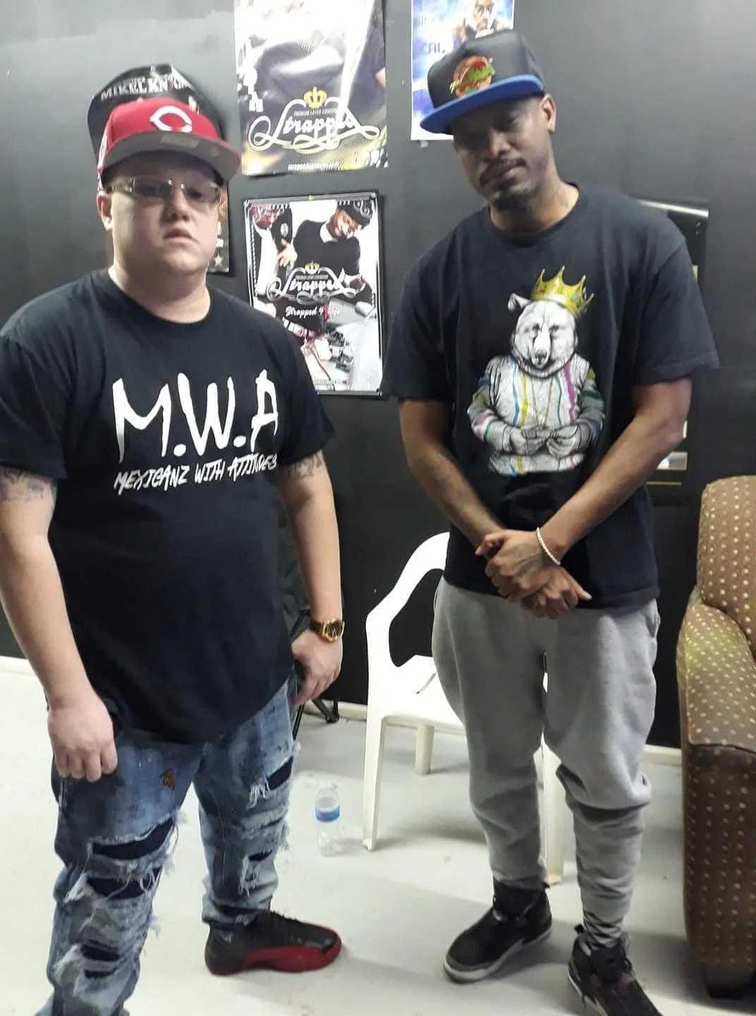 Two men standing indoors in front of black walls with posters. The man on the left wears a red and gray baseball cap, sunglasses, a black T-shirt with white lettering, distressed jeans, and black sneakers. The man on the right wears a black baseball 