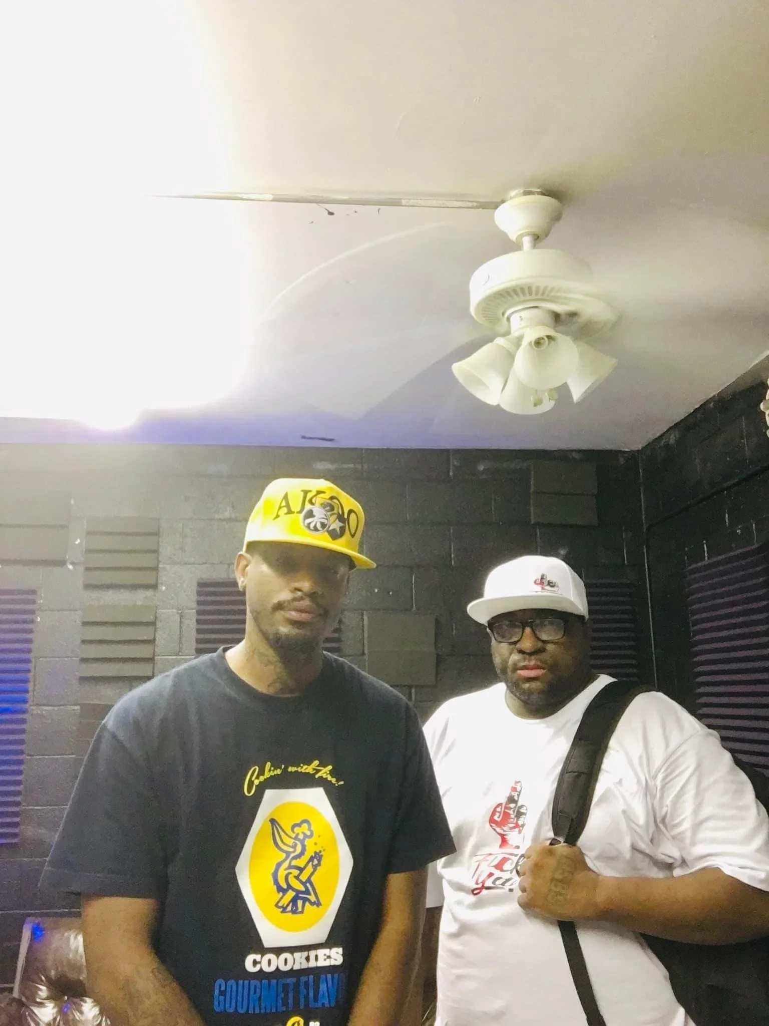 Two men posing in a recording studio with soundproofing foam on the walls, one wearing a yellow cap and black T-shirt, the other wearing a white cap, glasses, and a white T-shirt, both looking at the camera.