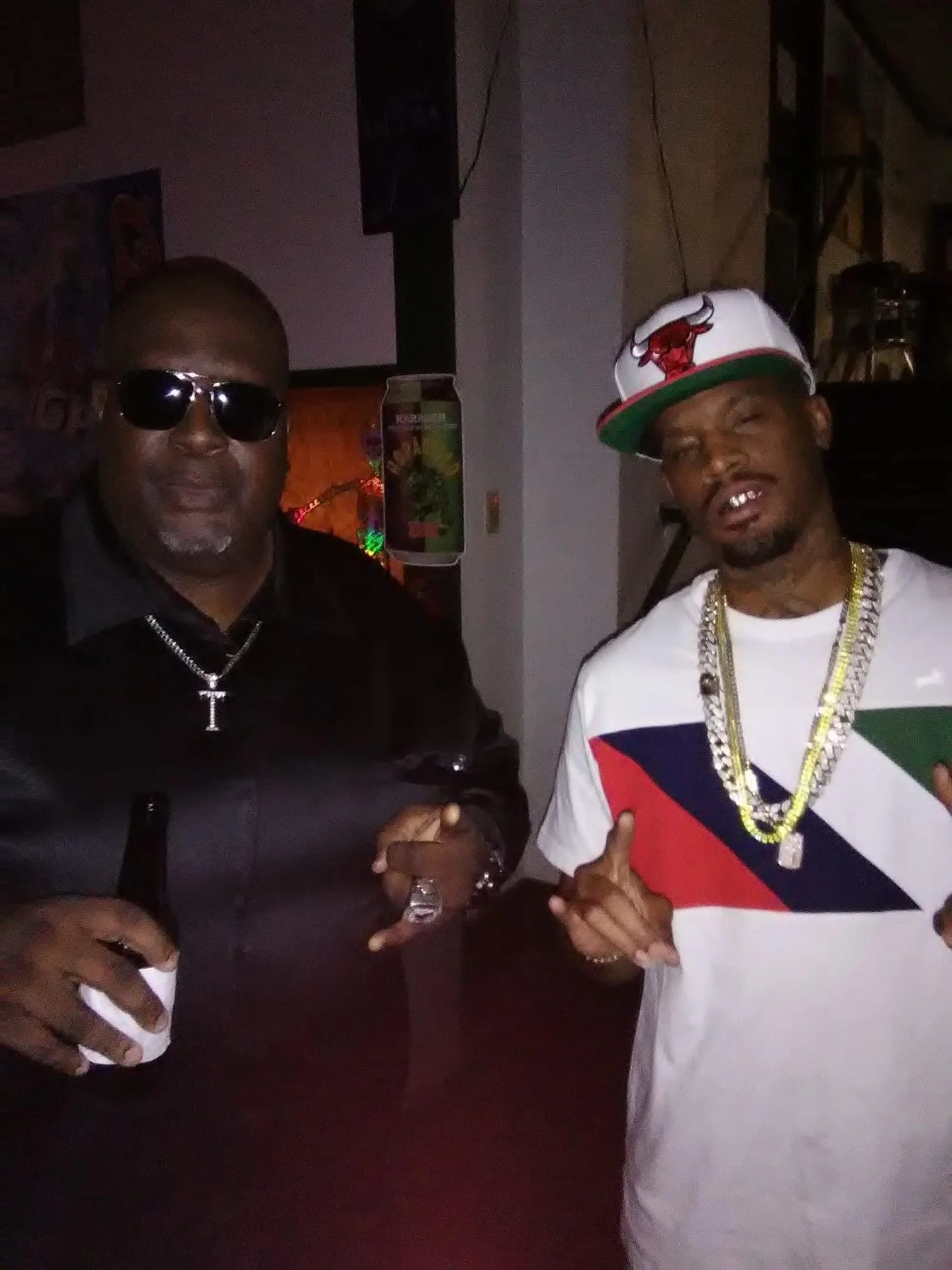 Two men posing for a photo indoors. The man on the left wears sunglasses, a black shirt, and has a chain with a cross pendant. The man on the right wears a white t-shirt with colored stripes, a cap with a red and white logo, and gold chains. Both are
