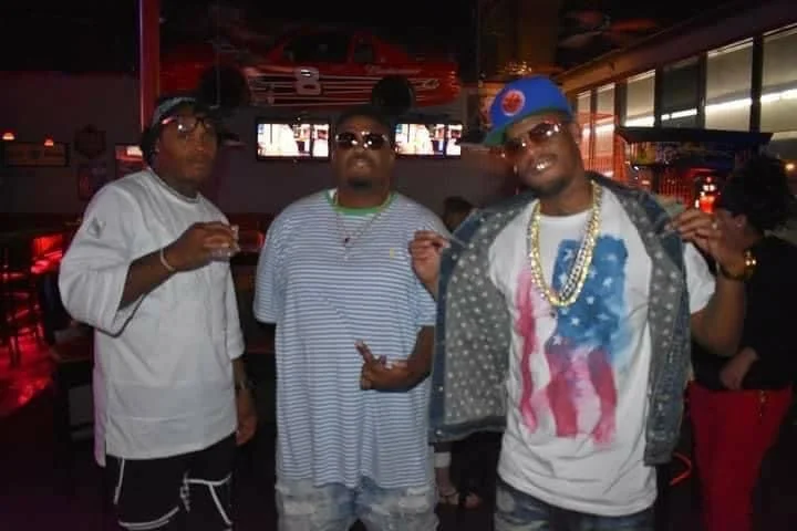 Three young men standing in a dimly lit bar or nightclub, dressed in casual hip-hop style clothing with chains and sunglasses, with one holding a drink.