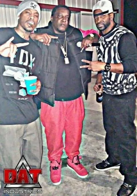 Three men posing together in a casual indoor setting, with two of them making hand gestures and holding drinks, one wearing a striped shirt, baseball cap, sunglasses, and black sneakers, the second wearing a hoodie, glasses, and black sneakers, and t