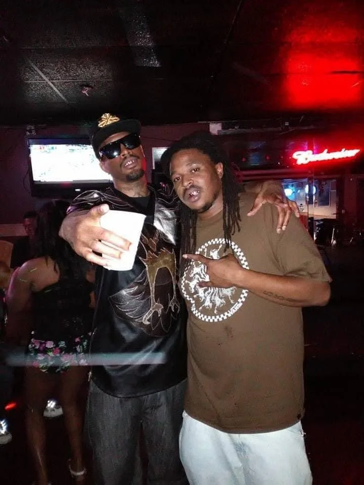 Two men posing together in a bar, one wearing sunglasses and a cap, and the other with dreadlocks, both making hand gestures.