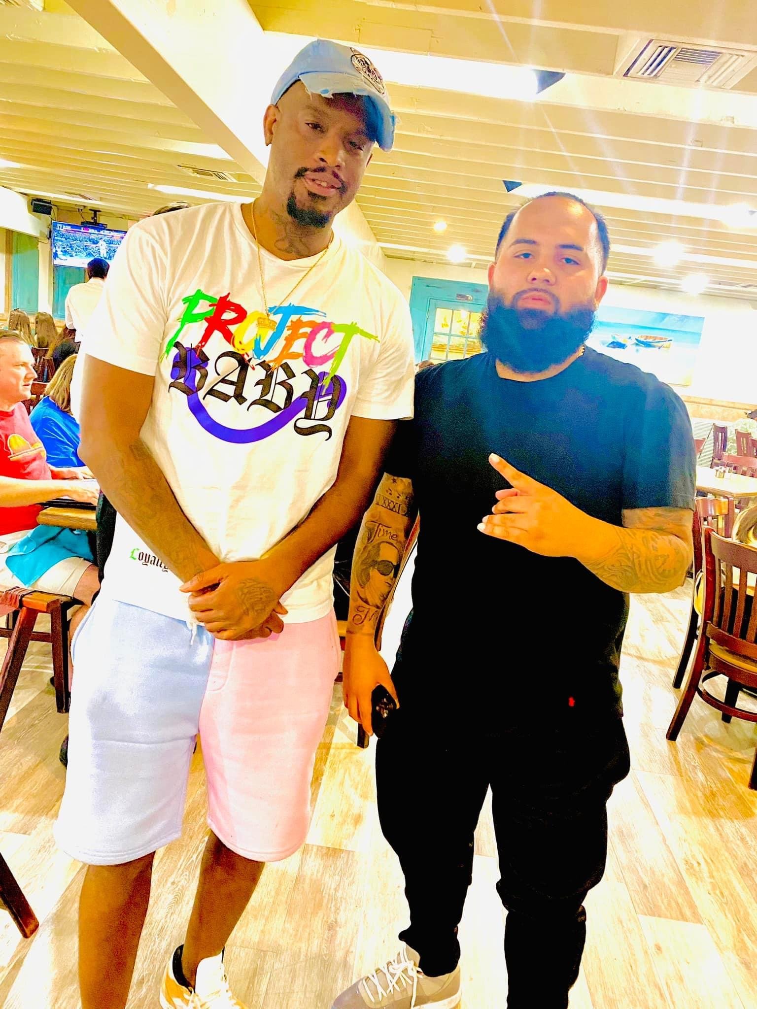 Two men standing together in a restaurant, one wearing a white t-shirt with colorful text and pink shorts, the other wearing a black t-shirt and black pants, both with tattoos and looking at the camera.