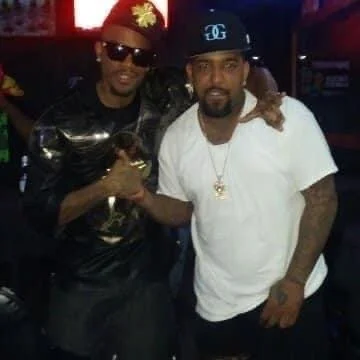 Two men standing together in a dimly lit setting, posing for the camera. The man on the left is wearing sunglasses, a red hat, and a leather jacket, while the man on the right is dressed in a white t-shirt and a black cap.