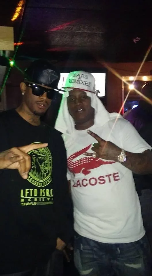 Two men posing together in a dimly lit indoor setting. The man on the left wears sunglasses, a black cap, and a black shirt with green graphics. The man on the right wears a white Lacoste t-shirt with a red crocodile logo, a white cap with green writ