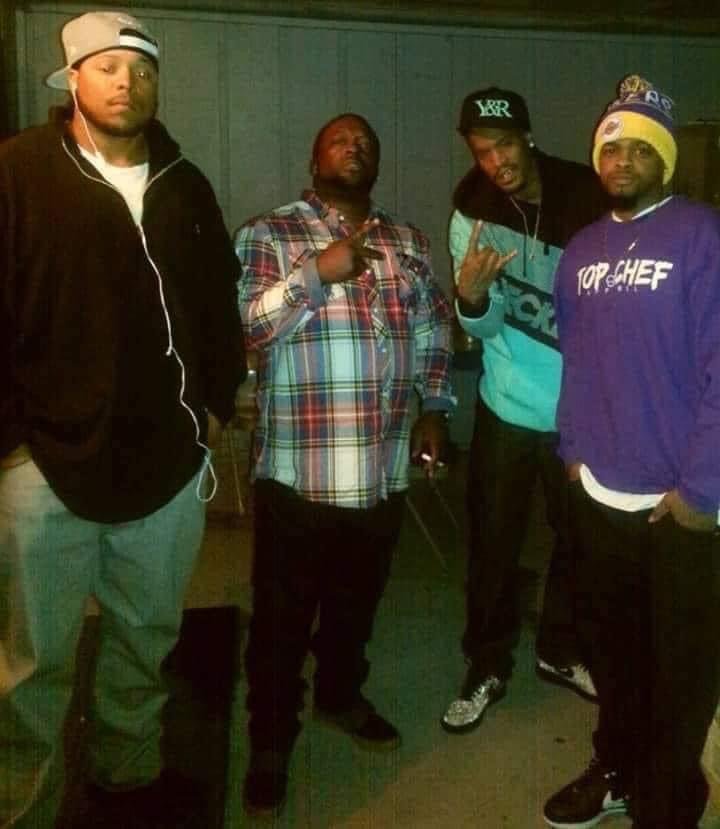 Four young men standing together in a casual indoor setting. One is wearing a black jacket, gray pants, and a backward cap. The second, in the middle, is wearing a plaid shirt and holding a cigarette. The third, to the right of the middle, is wearing