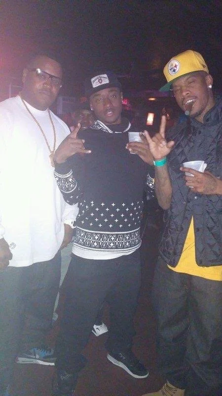 Three men posing together at a party or club, making hand gestures and holding cups, with dim lighting and a dark background.