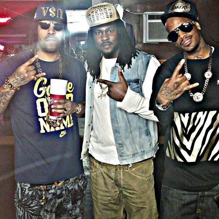 Three men with tattoos and jewelry posing indoors. The man on the left is wearing sunglasses, a cap, and a black Nike t-shirt with gold lettering, holding a red cup. The man in the middle has dreadlocks, a denim vest, and is pointing with his right h