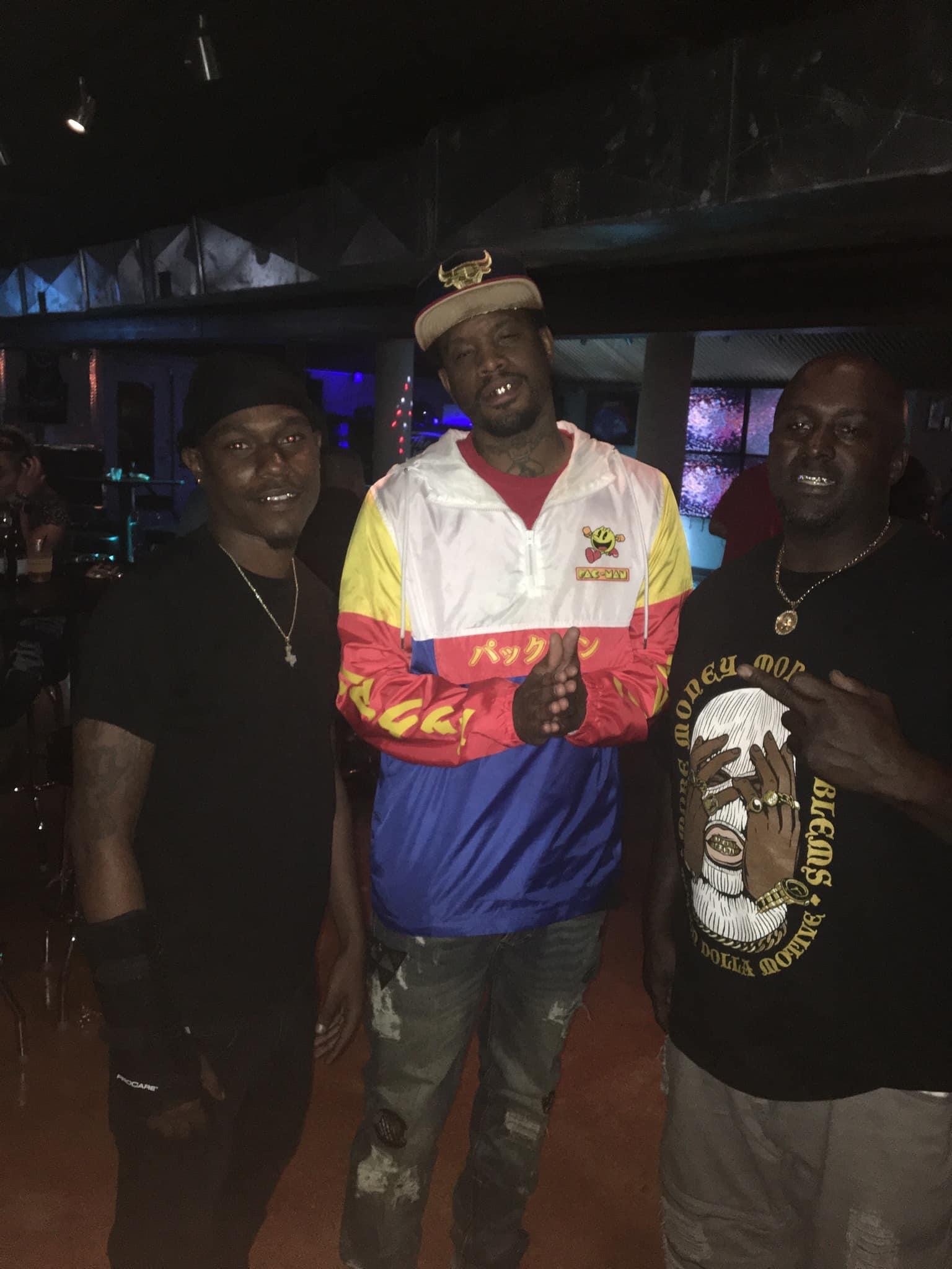 Three men standing together in a dimly lit indoor venue, with the man in the middle wearing a colorful jacket and a white cap with a bulls logo, flanked by two men in dark clothing.
