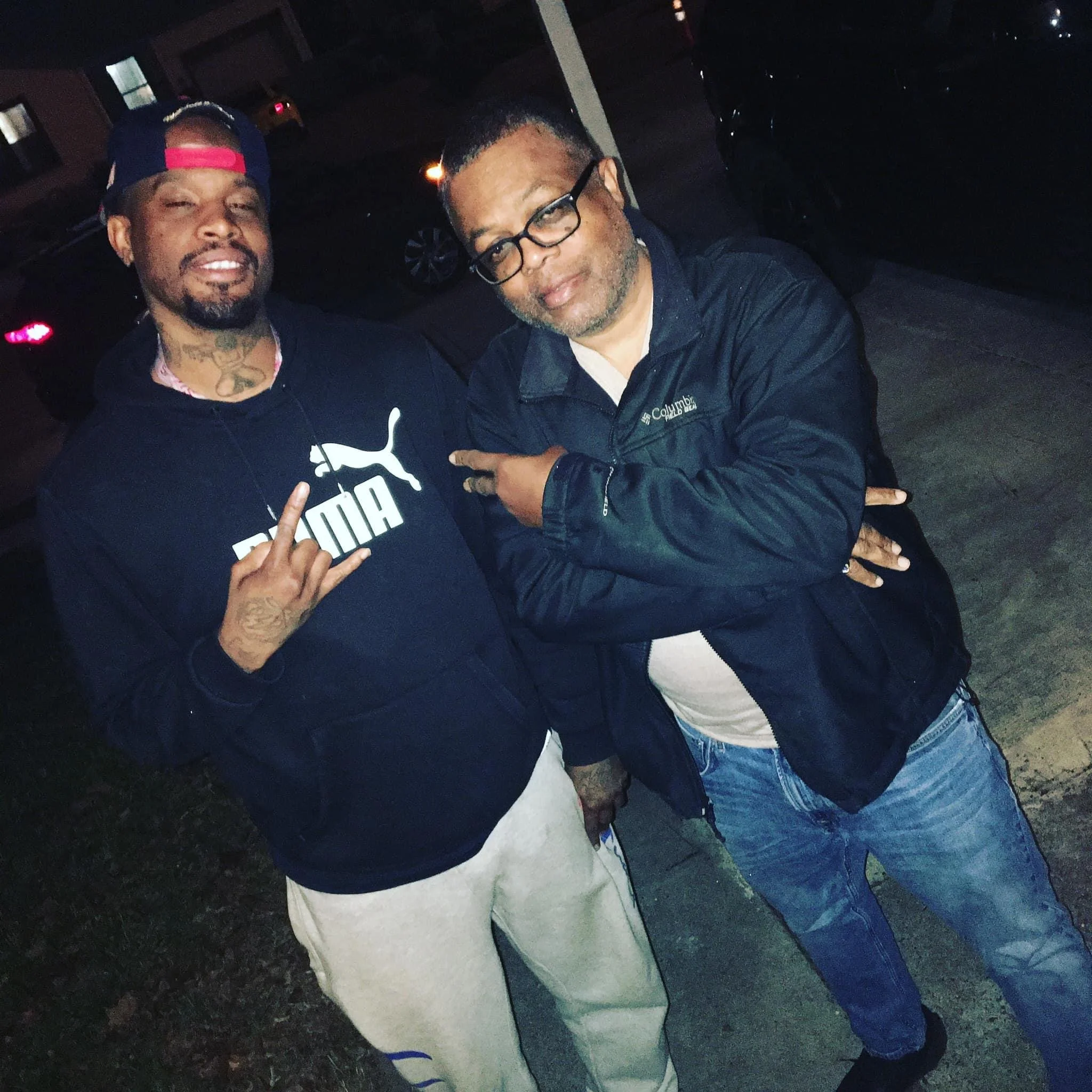 Two men standing outdoors at night, one wearing a Puma hoodie and making a hand gesture, the other pointing at him with his arms crossed, both smiling.