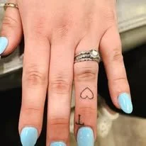 Close-up of a hand with a heart and the letter 'L' tattooed on one finger, and a wedding ring on the ring finger. The person has light blue nail polish.