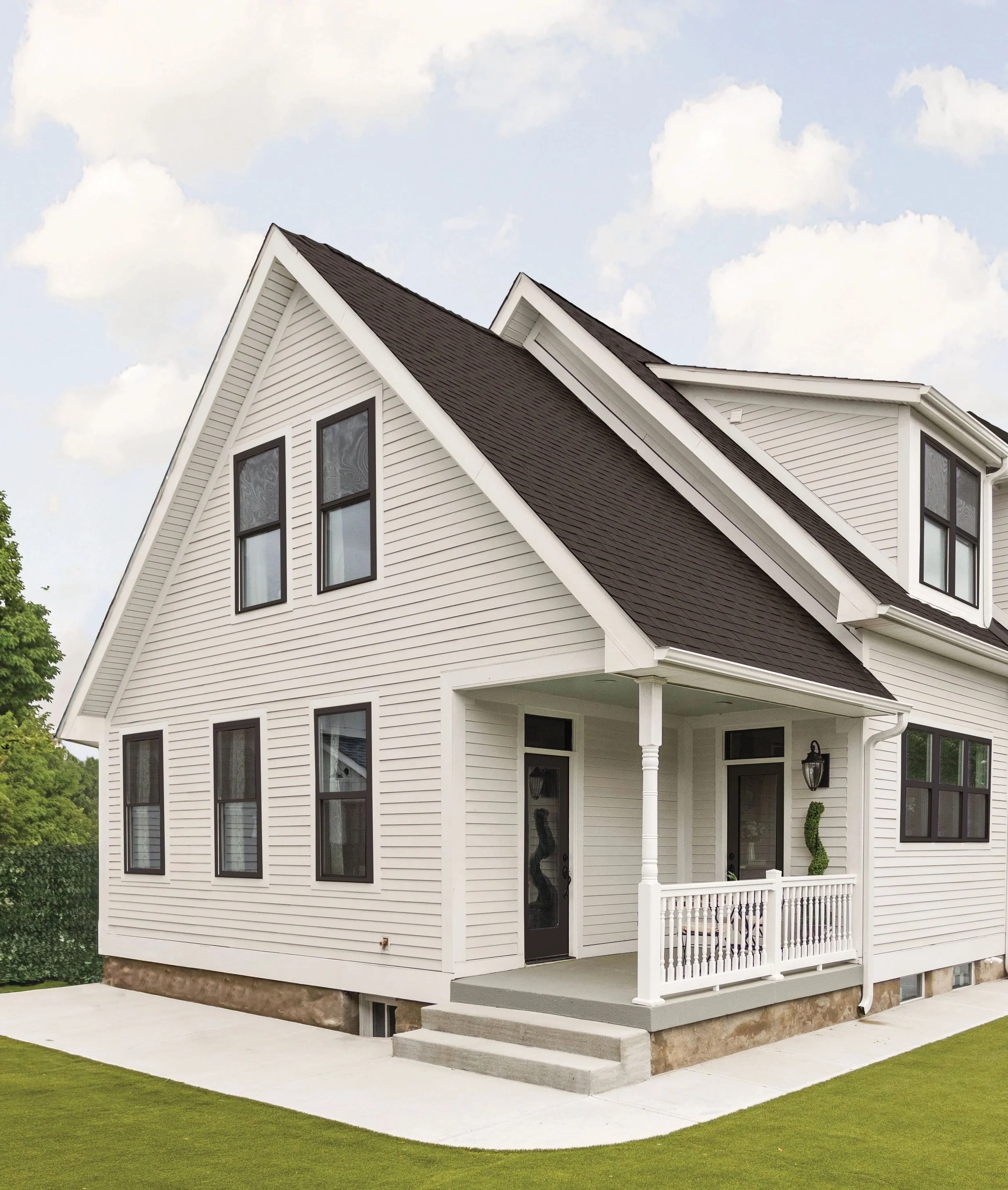 White two-story house with black windows, a front porch, and a gabled roof, surrounded by a green lawn and a sidewalk under a partly cloudy sky.