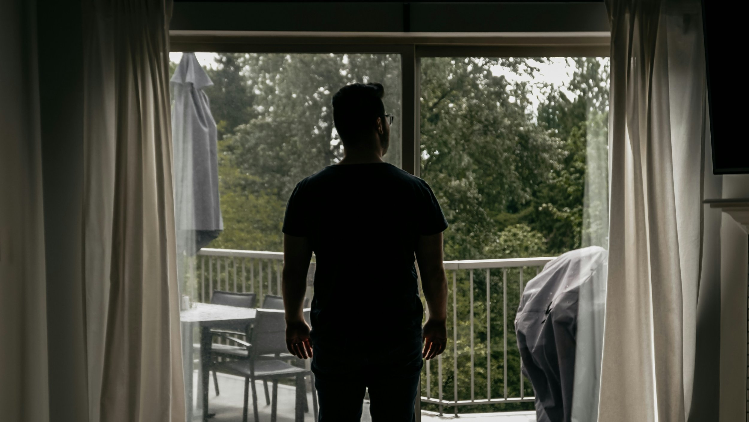 A silhouette of a man standing inside near a sliding glass door, looking out onto a balcony with trees and outdoor furniture.