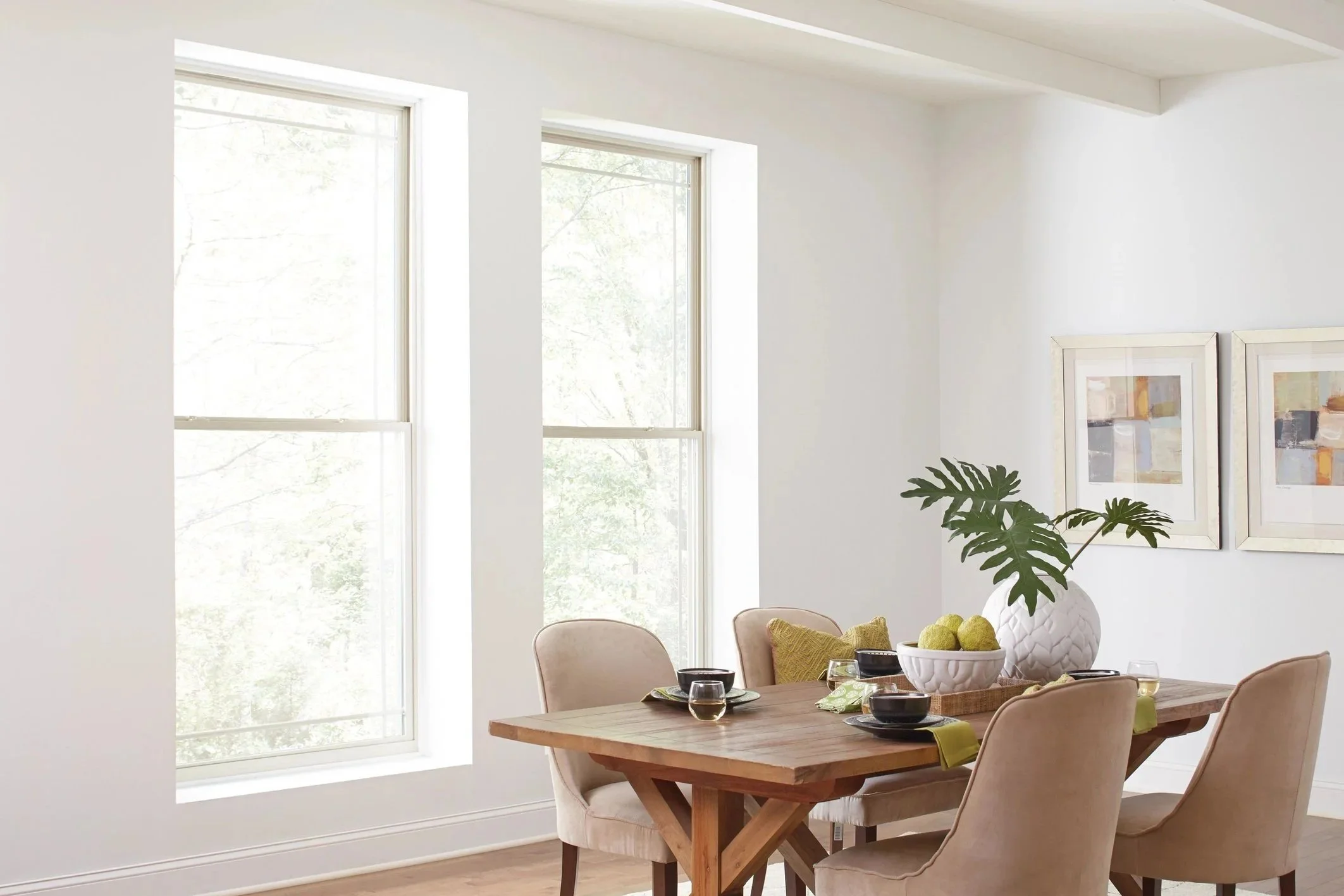 A dining room with a wooden table set for four, decorated with a large white vase containing a leafy green plant and a bowl of green fruits. There are four beige upholstered chairs around the table, and the room has white walls with two framed abstract artworks. Large windows allow natural light to fill the space.