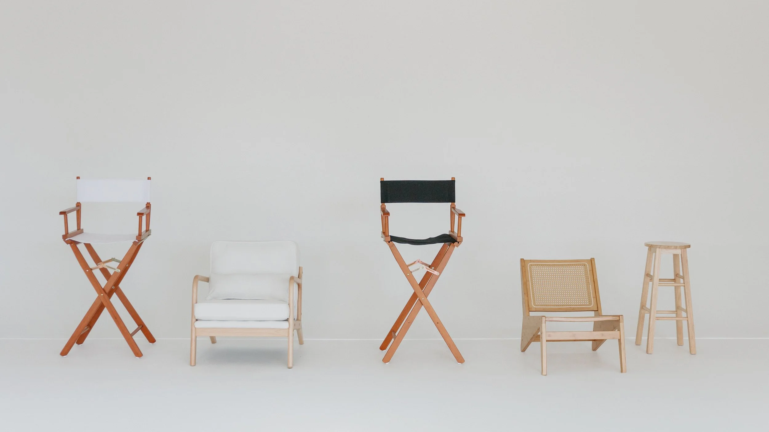 Various chairs and stools arranged in a row against a plain white wall, including a director's chair, a white armchair, a black director's chair, a woven rattan chair, and wooden stools.