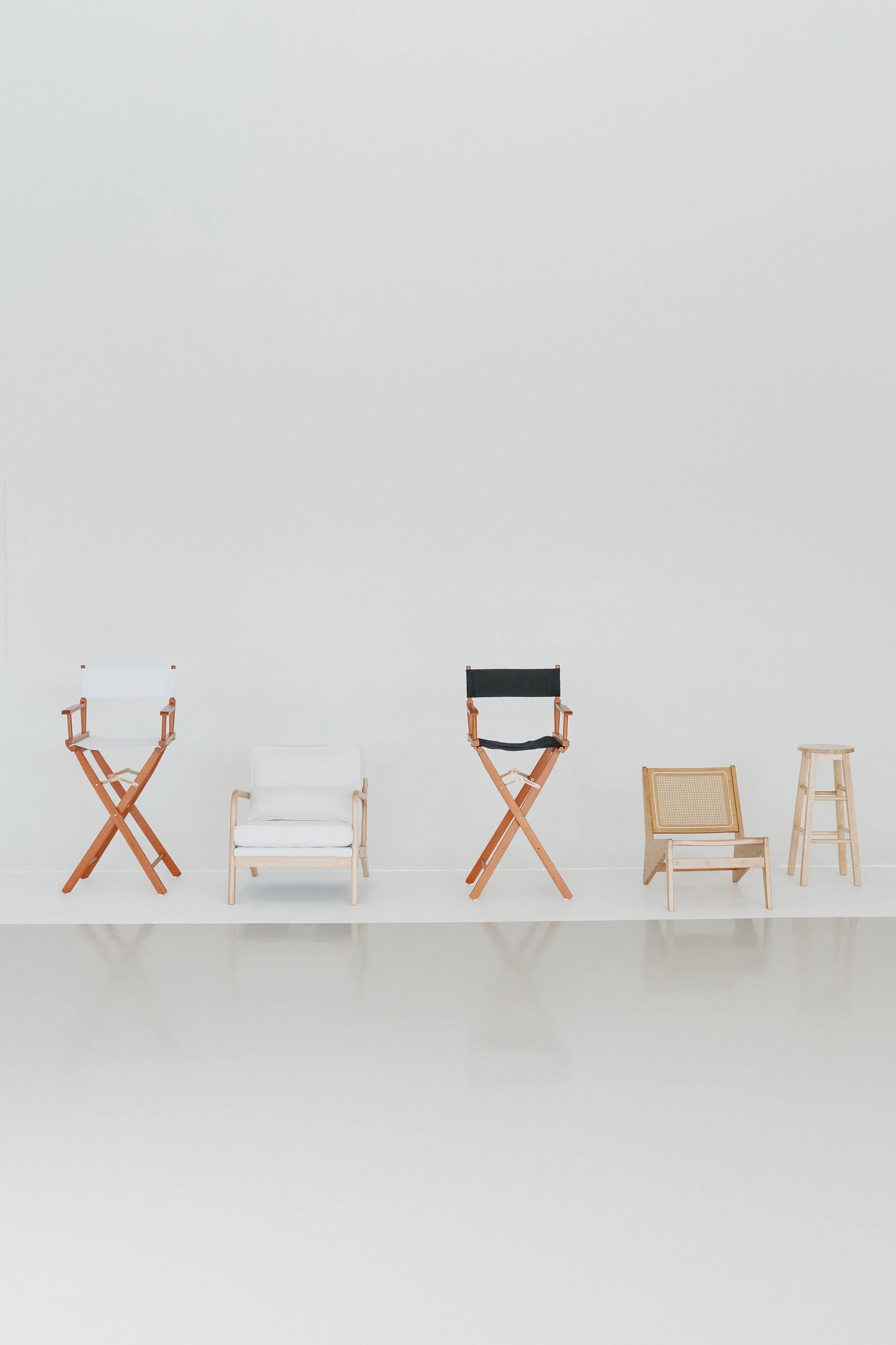 A plain white wall with a row of various chairs and stools arranged on the floor in front of it.