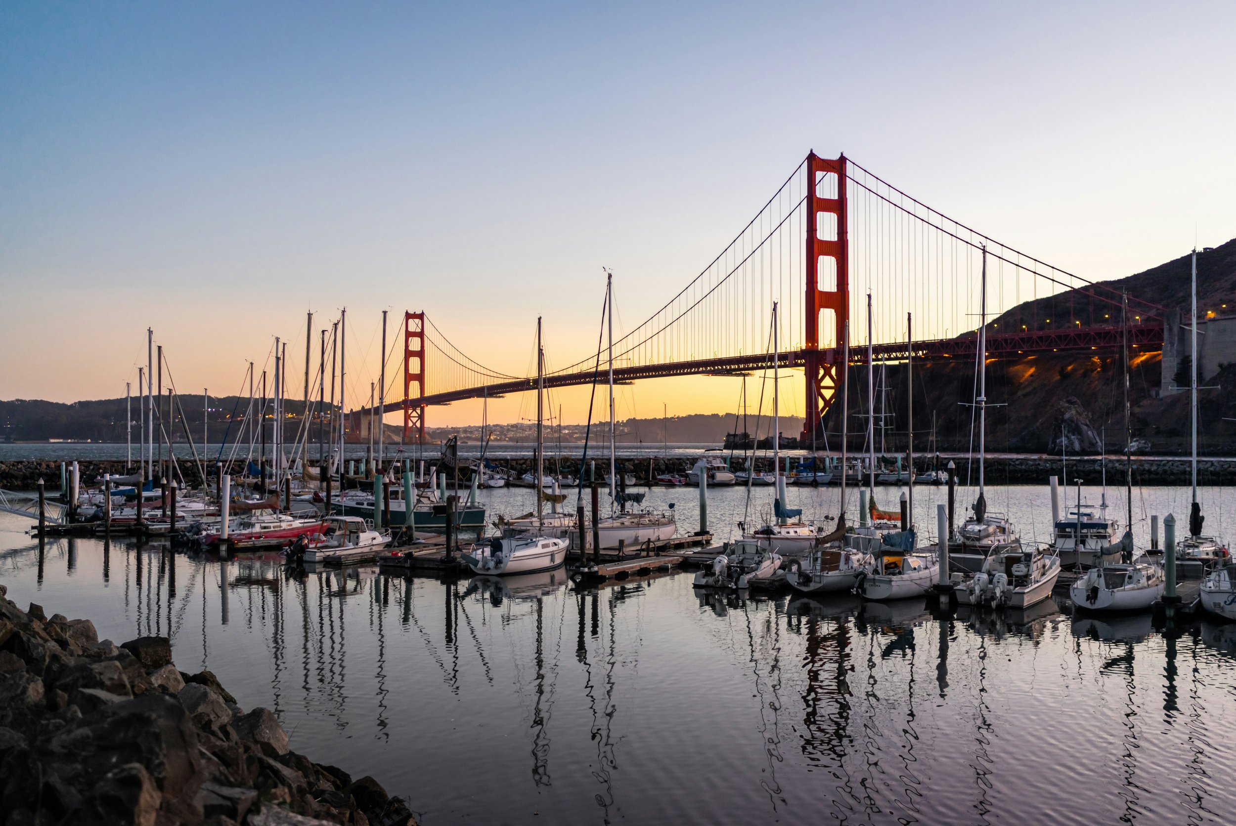 A marina with boats docked, the Golden Gate Bridge in the background at sunset, and hills on the horizon.