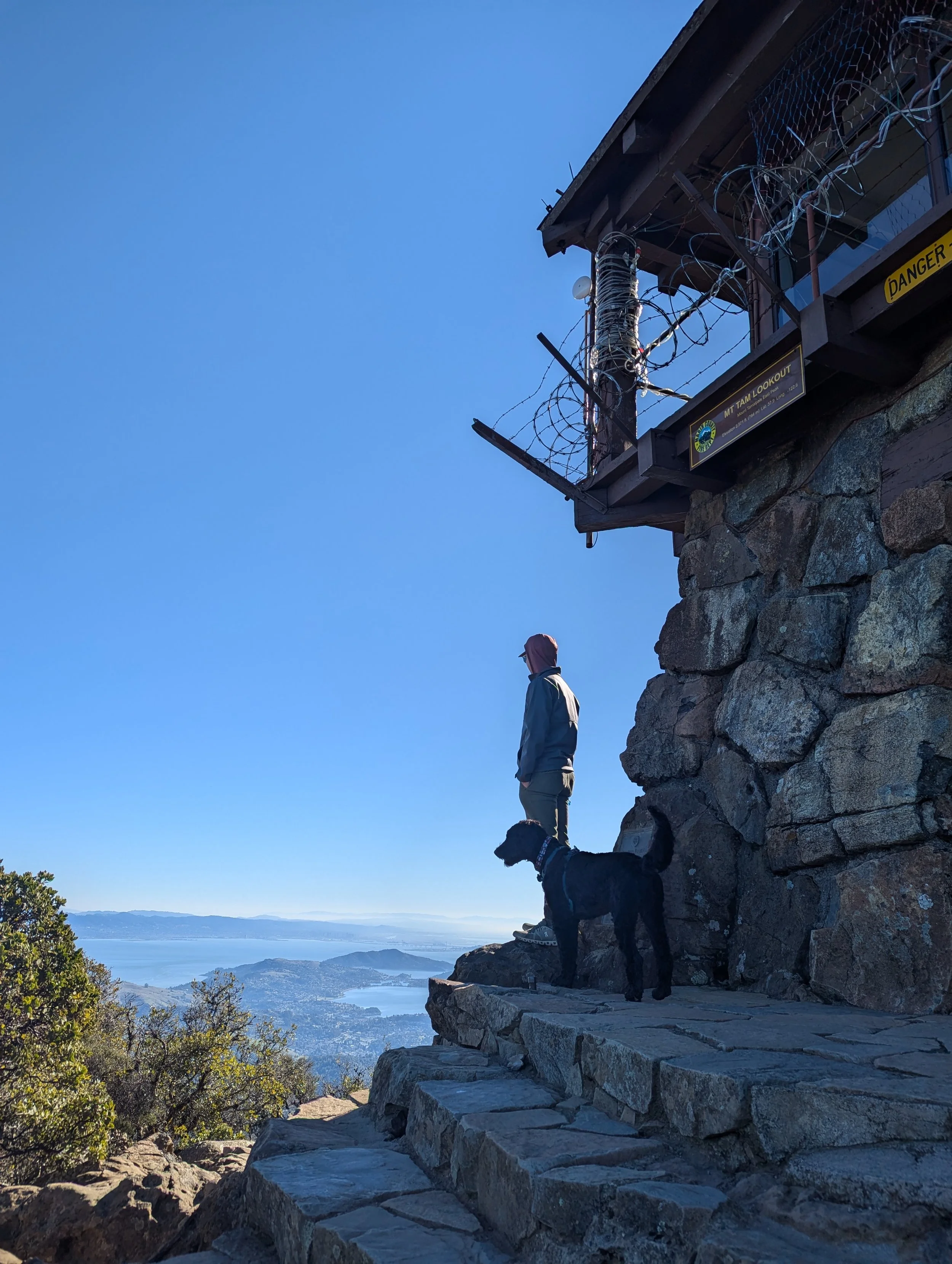 A person with a pink head covering and light-colored jacket standing on stone steps overlooking a large body of water and distant mountains, with a black and white dog standing on the steps beside them. The scene is set outdoors during the day, with a clear blue sky.