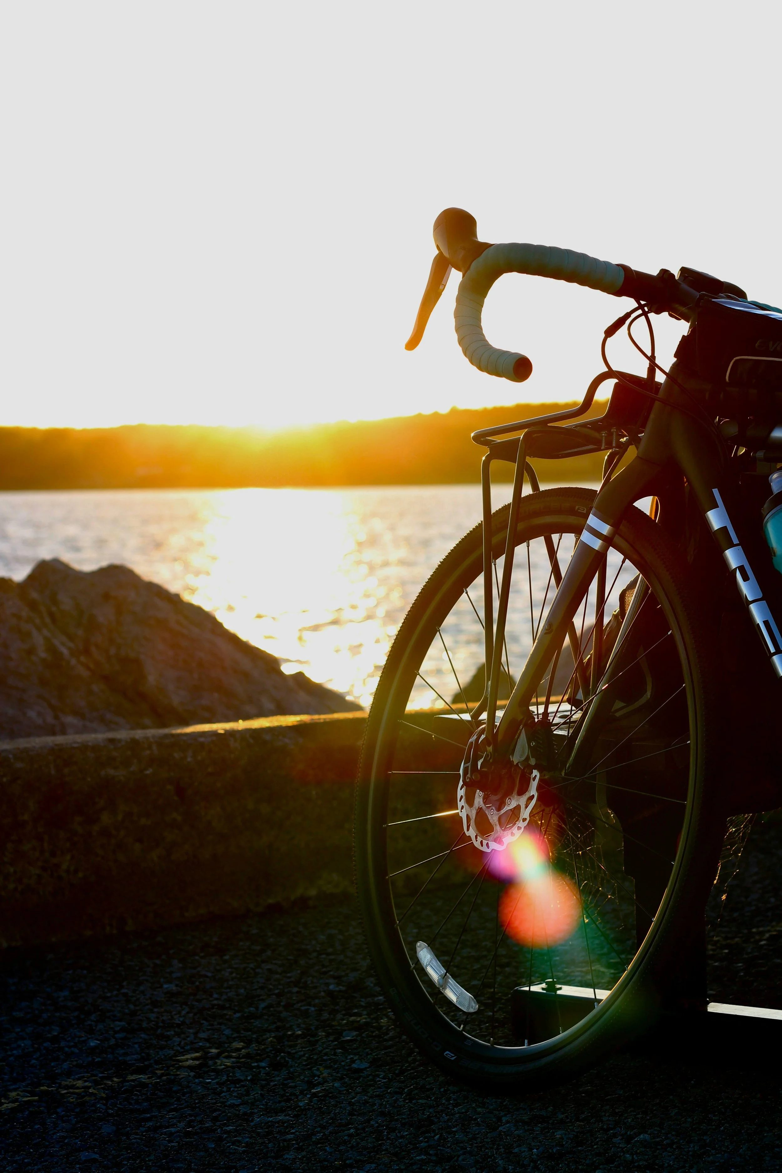 A bicycle parked on a dark paved path near water, with a sunset or sunrise in the background casting a golden glow, and the bicycle's handlebars and front wheel visible in the foreground.
