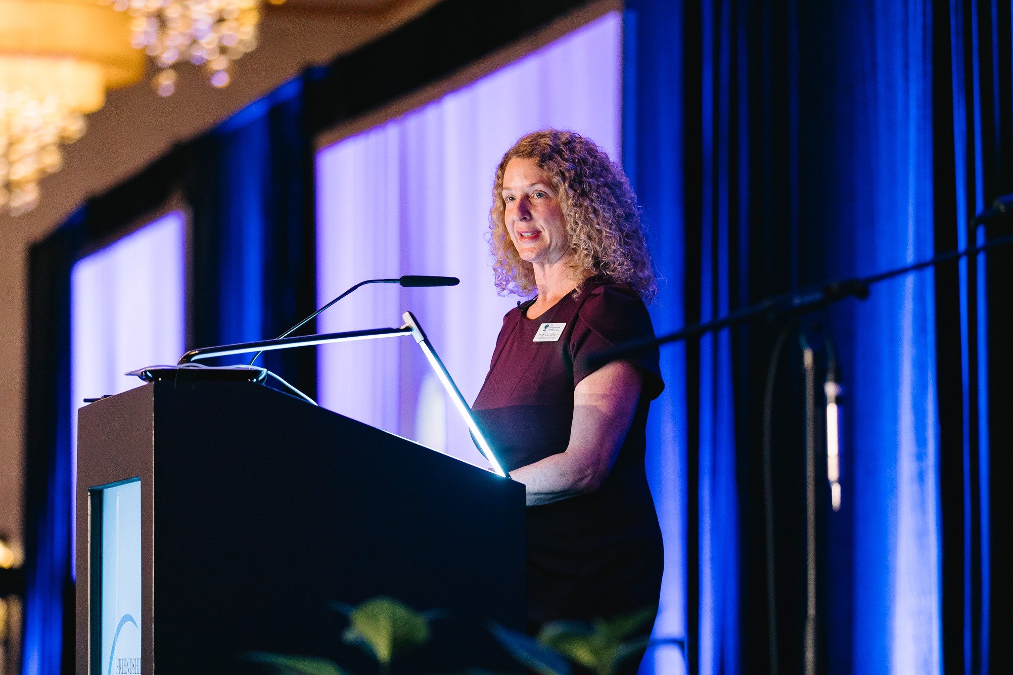 A woman with curly blonde hair giving a speech at a podium during an event with blue lighting and draped curtains in the background.