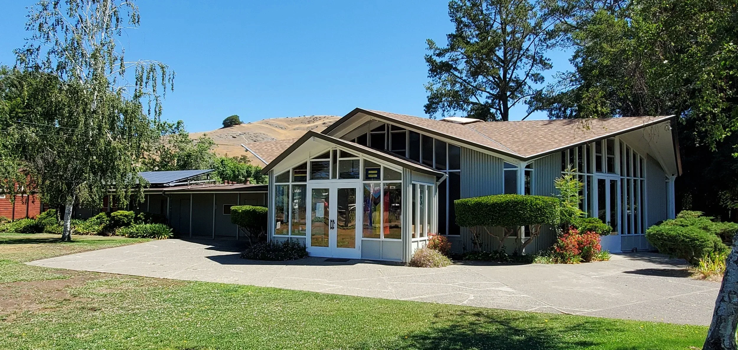 Christ Presbyterian Church - a modern building with large glass windows surrounded by green bushes and trees, under a blue sky with a hillside in the background.