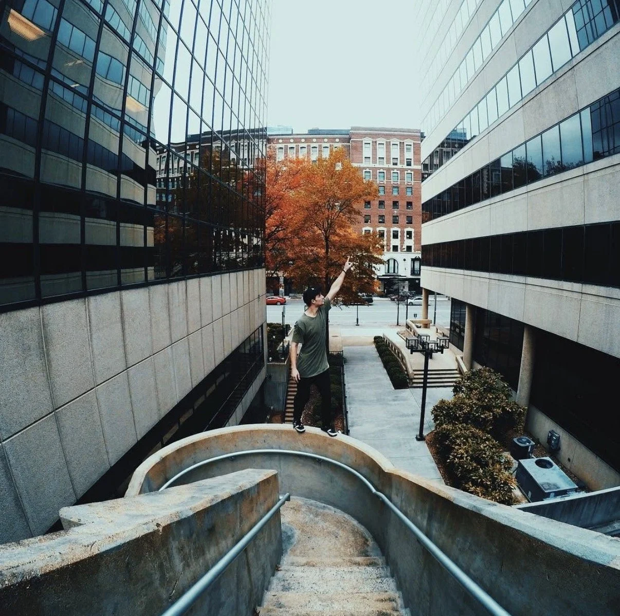 A person standing at the top of a concrete staircase between two modern office buildings, pointing upwards towards the sky, with a tree with orange autumn leaves and urban buildings visible in the background.