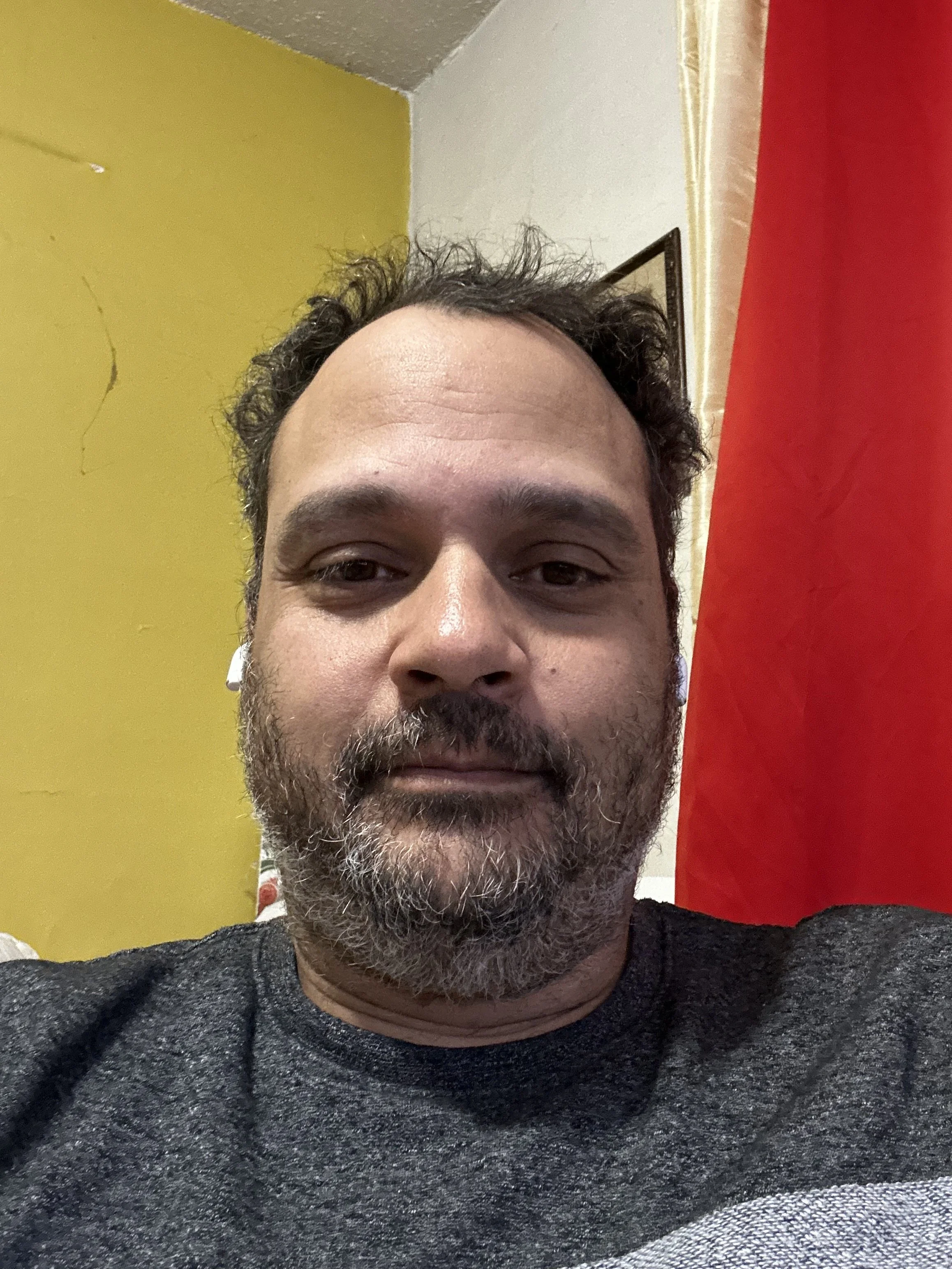Close-up of a man with dark curly hair, a beard with some gray, wearing a dark gray shirt and white earphones, sitting indoors in front of a yellow wall with a cracks and a red curtain.