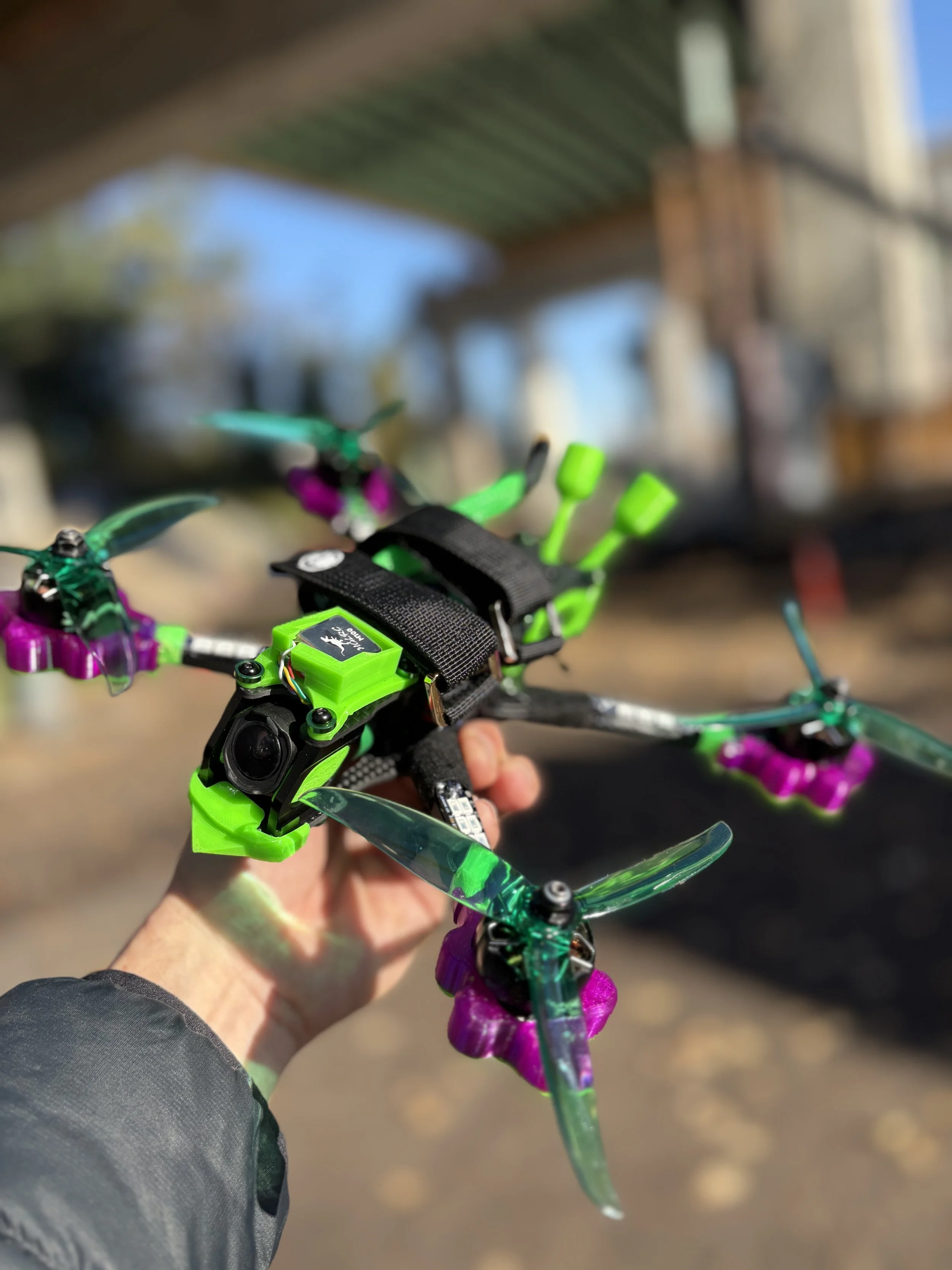 A person holding a colorful drone with purple and green accents outdoors on a sunny day, with blurred background of a house and trees.