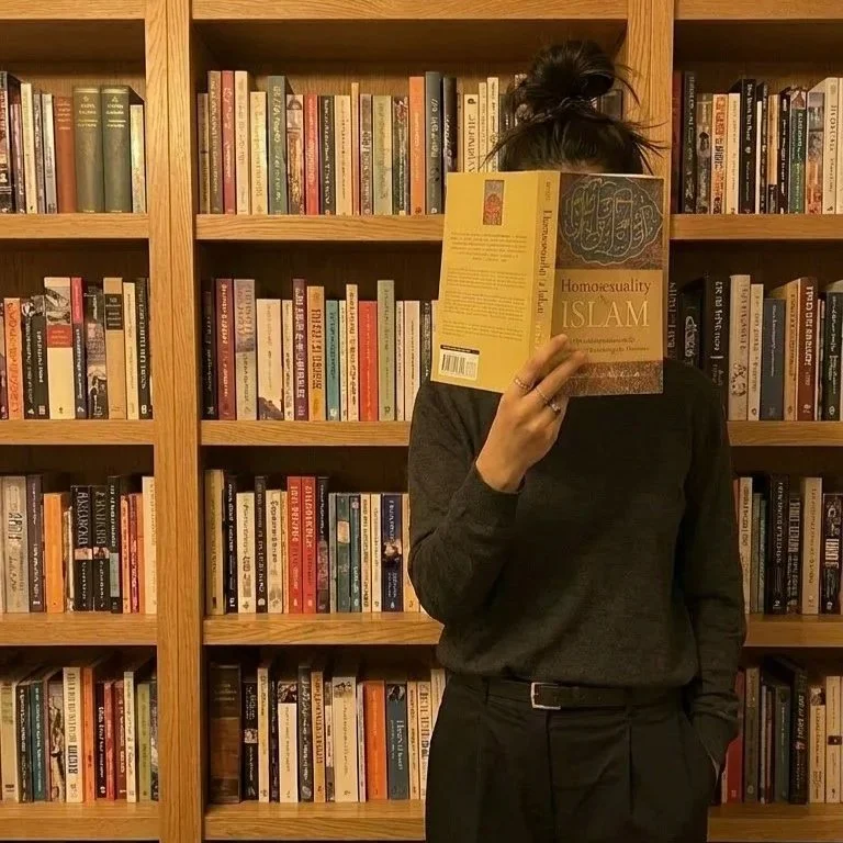 Person standing in front of a bookshelf, holding a book titled 'Homosexuality & Islam' in front of their face, wearing a dark long-sleeve top and with hair tied up in a messy bun.